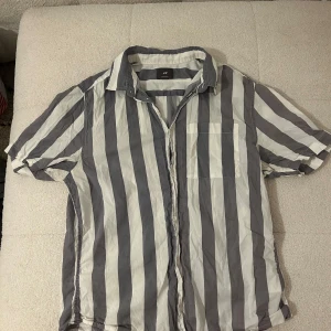 Randig kortärmad skjorta från H&M - Snygg kortärmad skjorta från H&M i vitt och  blå/grått med breda vertikala ränder. Skjortan har klassisk krage, bröstficka och knäppning framtill. 