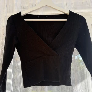 Svart omlottopp med v-ringning - Säljer en svart omlottopp med v-ringning och långa ärmar. Toppen är ribbad och har en croppad passform som ger en snygg siluett. Perfekt att styla med höga jeans eller kjol för en trendig look.