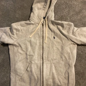Grå hoodie från Polo Ralph Lauren - Snygg ljusgrå hoodie från Polo Ralph Lauren med dragkedja, huva och snörning. Klassisk broderad logga på bröstet och två fickor framtill. Perfekt för chill dagar och enkel att matcha med jeans eller joggers.