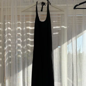 Svart långklänning från H&M - Svart långklänning från H&M med smala axelband som knyts i nacken. Klänningen har en djup urringning fram och är ärmlös, vilket ger en clean och stilren look. Perfekt för dig som gillar minimalistisk stil och vill ha något enkelt men snyggt.