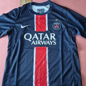 PSG Nike Kit - Paris Saint-Germain fotbollströja från Nike i marinblått med en bred röd vertikal rand på framsidan och vita detaljer. PSG-logga med guldstjärna på bröstet, Qatar Airways tryck och klassisk rund hals. Tillverkad i lätt, ventilerande polyester för maxad komfort på planen.