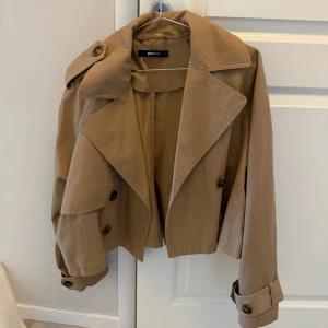 Croppad beige trenchcoat  - Säljer en croppad trenchcoat i beige/ljusbrun förg från Gina Tricot. 