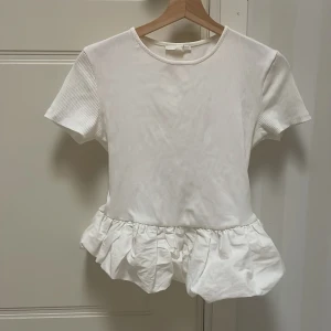 Vit blus med volangkant - Supersöt vit blus med korta ärmar och rund hals. Toppen har ribbade partier upptill och en bred volangkant nedtill som ger en lekfull och trendig look. Perfekt att styla med jeans eller kjol för en fräsch outfit. Köpt på Zara i Paris, lite för kort. Annars jätte fin topp. 