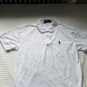 Vit polo t-shirt från Ralph Lauren - Fräsch vit polo t-shirt från Ralph Lauren med klassisk krage och två knappar framtill. Ikoniska broderade loggan i marinblått på bröstet. T-shirten är kortärmad och tillverkad i mjuk bomull som andas. Perfekt för en clean och stilren look.