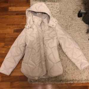 Beige quiltad jacka - Säljer en beige quiltad pufferjacka från H&M med avtagbar huva och snygga vågiga sömmar. Jackan har knappar framtill, två fickor och en lite oversized passform. Perfekt för kyliga dagar och enkel att styla till jeans eller leggings💗💗