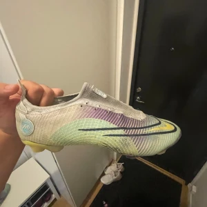 Nike Mercurial fotbollsskor i pastell - Nike Mercurial fotbollsskor med en futuristisk design i pastellfärger som lila, gul, grön och silver. Skorna har en låg profil, rund tå och platt sula med dobbar för gräsplan. Ovandelen är i syntetmaterial med diskret struktur och stora Swoosh-loggor på sidorna.