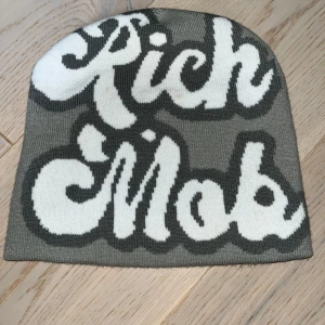 Grå mössa med Rich Mob-tryck - Säljer en grå mössa från Rich Mob med stor, vit text i coolt grafiskt typsnitt på framsidan. Mössan är stickad och har en enkel, rak form som sitter snyggt på huvudet. Perfekt för dig som gillar streetwear och vill sticka ut med ett statement.