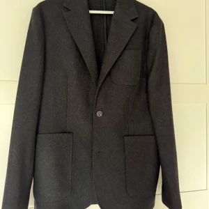 Mörkgrå ullkavaj  från Lexington  - Stilren mörkgrå kavaj från Lexington i storlek M. Klassisk modell med tre utanpåfickor, enkelknäppt framtill och mjuk känsla i ullmaterial. Perfekt för höst plagg