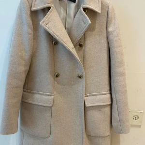  Ny Beige skepparkavaj från Zara - Stilren beige skepparkavaj från Zara med dubbelknäppning och stora fickor framtill. Jackan har klassisk krage och är helfodrad, perfekt för dig som vill ha en clean och tidlös look. Materialet är mjukt och känns lyxigt.