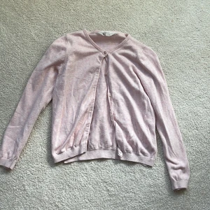 Ljusrosa kofta från H&M - Söt ljusrosa kofta från H&M med v-ringning och knappar framtill. Långärmad modell i mjuk bomullsblandning, perfekt för lager på lager. Ribbstickade muddar vid ärmslut och nederkant ger en snygg passform.