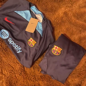 FC Barcelona Nike träningströja marinblå - Snygg FC Barcelona träningströja från Nike i marinblått med ljusblå detaljer. Tröjan har klubbmärke på bröstet, röd Nike-logga och Spotify-tryck. Materialet är lätt och andas, perfekt för fotbollsträning. Halv dragkedja framtill och sportig passform.