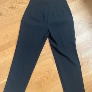 Svarta högmidjade kostymbyxor - Snygga svarta kostymbyxor med hög midja och raka ben. Byxorna har pressveck och diskreta fickor bak, vilket ger en clean och stilren look. Perfekta för dig som gillar en klassisk men modern vibe.