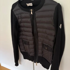 Svart kofta från Moncler med dragkedja - Svart kofta från Moncler med quiltad framsida och två fickor med knapp. Populär & petfekt nu till hösten!! Den har lite slitningar (se bilder) men det är inget man tänker på när den används. 