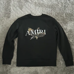 Svart sweatshirt från Amiri med örn - Svart sweatshirt från Amiri med stort tryck av en örn och AMIRI-logga på bröstet. Tröjan har rund hals, långa ärmar och ribbade muddar. Perfekt för dig som gillar streetwear och vill sticka ut med en snygg detalj framtill. S men sitter som en liten XS / barnstorlek (passar tca 165–170 cm). Väldigt bra skick, används några gånger. 