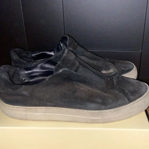 Svarta sneakers från Axel Arigato - Svarta slip-on sneakers från Axel Arigato med minimalistisk design och vit platt sula. Skorna har diskret logga på sidan och är tillverkade i mocka med insida i skinn. Perfekta för en clean och stilren look.