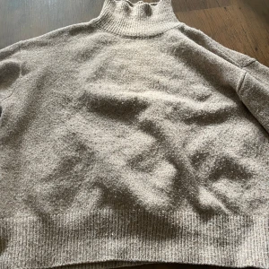 Beige stickad polotröja - Beige stickad polotröja med ribbade muddar och hög krage. Tröjan har en loose fit och är tillverkad i ett mjukt ullmaterial som känns riktigt cozy. Perfekt för kyliga dagar när du vill ha något både snyggt och bekvämt.