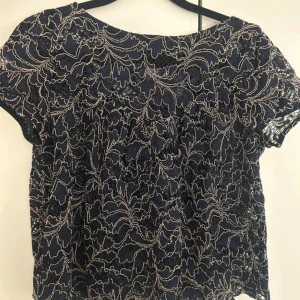 Mörkblå spetsblus från Zara Basic - Superfin mörkblå blus från Zara Basic med korta ärmar och genombruten spets. Blusen har ett broderat mönster i beige och en luftig, lätt form. Perfekt för dig som gillar detaljer och vill sticka ut lite extra.