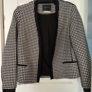 Svartvit rutig kavaj Maison Scotch - Snygg svartvit kavaj från Maison Scotch med klassiskt hundtandsmönster. Kavajen har lång ärm, svarta detaljer vid fickorna och en öppen front utan knäppning. Perfekt för dig som vill sticka ut med en tidlös och trendig look.