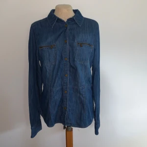 Jeansskjorta från Peak Performance - Snygg jeansskjorta från Peak Performance i klassisk blå denim. Skjortan har långa ärmar, tryckknappar framtill och två bröstfickor med dragkedja. Broderad logga på sidan ger en extra detalj. 