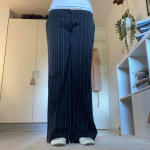 Svarta kostymbyxor med vita ränder - Snygga svarta kostymbyxor med vita tunna ränder i ett klassiskt randigt mönster. Byxorna har låg midja, raka vida ben och hällor för bälte. Perfekta för en stilren och trendig look. Har sytt upp byxorna då de var för långa för mig som är 170 cm.