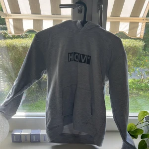 Grå Hov1 hoodie med tryck - Säljer en grå hoodie från Hov1 med svart tryck på bröstet och texten 'Hon dansar vidare i livet..' på ryggen. Tröjan har huva, magficka och är i mjuk bomullsblandning. Perfekt för dig som gillar svensk musik och vill ha en chill stil.