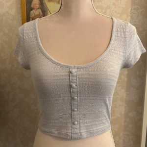 Ljusblå croppad topp från Primark - Supersöt ljusblå croppad topp från Primark med korta ärmar och snygg mönstrad struktur. Djup rund halsringning och dekorativa knappar framtill, i storlek 2XS. 