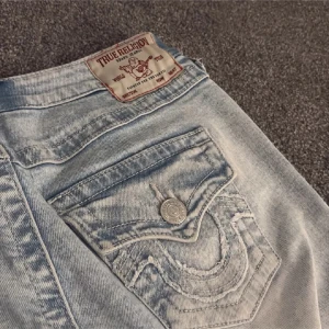 Ljusblå True Religion jeans - Säljer ett par ljusblå jeans från True Religion med ikonisk bakficka och snygga kontrastsömmar. Jeansen har klassisk femficksdesign och cool tvättad look. Perfekta för dig som gillar streetwear och vill sticka ut.