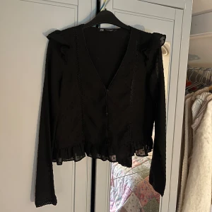 Svart prickig blus med volang från Zara - Supersnygg svart blus från Zara med diskret prickigt mönster, v-ringning och knappar framtill. Långa ärmar med volangdetaljer vid axlarna och nederkant. Blusen har transparenta partier och en söt volang längst ner som ger extra stilpoäng.