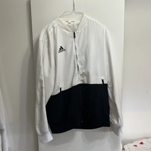Vit och svart bomberjacka från Adidas - Snygg bomberjacka från Adidas i vit och svart färgblock-design. Jackan har dragkedja framtill, två fickor och Adidas-logga på bröstet. Tillverkad i polyester med lång ärm och klassisk bomberkrage. Perfekt för en sportig och clean stil.