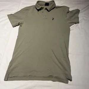 Grön Peak Performance pikétröja - Snygg grön pikétröja från Peak Performance med klassisk krage och knappslå framtill. T-shirten har korta ärmar och en liten broderad logga på bröstet. Tillverkad i mjuk bomull som känns skön mot huden. Perfekt för en chill och stilren look.