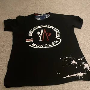 Svart Moncler t-shirt med tryck - Svart t-shirt från Moncler med stort grafiskt tryck och logga framtill i vitt och rött. Coola vita färgstänk nertill och broderad Moncler-patch på ärmen. Rund hals med insida i blått med serietidningsmönster. Passar dig som gillar streetstyle och exklusiva märken.