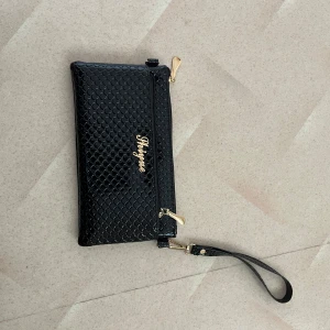 Svart kuvertväska från Shizjune - Snygg svart kuvertväska från Shizjune med glansig yta och fiskfjällsmönster. Väskan har guldfärgade detaljer, dragkedjor och en avtagbar handledsrem. Perfekt för att bära det viktigaste och ge din outfit en extra touch.