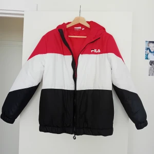 Pufferjacka från FILA XS - Snygg pufferjacka från FILA i storlek XS med färgblock i rött, vitt och svart. Jackan har huva, dragkedja framtill och elastiska muddar. Perfekt för dig som gillar sportig streetstyle och vill sticka ut med en cool look. Endast använd nån gång så I toppskick, notera att det finns ett litet svart märke på ärmen (kanske går att få bort).