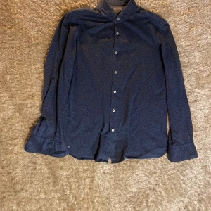 Mörkblå mönstrad skjorta från Xacus - Snygg mörkblå skjorta med diskret småmönster och klassisk krage. Skjortan har långa ärmar och knäppning framtill med vita knappar. Perfekt för dig som gillar stilrena detaljer och en bekväm passform. En liten fläck på baksidan av skjortan men den går bort med en tvätt.