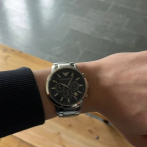 Emporio Armani silverfärgad klocka - Snygg klocka från Emporio Armani med svart urtavla och silverfärgat armband i metall. Klockan har kronograf-funktion och tydliga indexmarkeringar. Perfekt accessoar för dig som gillar stilrena detaljer och klassisk design. Köpte den tidigare detta år för 5400kr