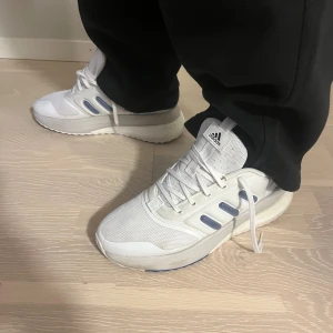 Vita Adidas sneakers med blå detaljer - Fräscha vita Adidas sneakers med blå detaljer och klassiska tre ränder på sidan. Ovandel i mesh och syntet, boost-sula för extra komfort och snörning framtill. Perfekta för dig som gillar sportiga skor, funkar jättebra till träning.