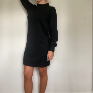 Svart stickad klänning från JDY - Mysig svart stickad klänning från JDY med långärmade ärmar och ribbad hög hals. Modellen är kort och har en loose passform som gör den superbekväm. Perfekt för dig som gillar enkel och stilren design med en touch av vardagschic.