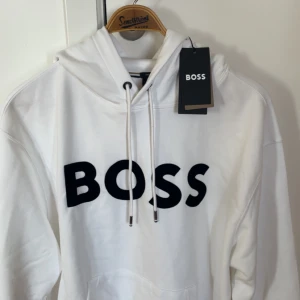 Vit oversized hoodie från BOSS - Säljer en helt oanvänd vit hoodie från BOSS med stor svart logga framtill. Tröjan är oversize. 