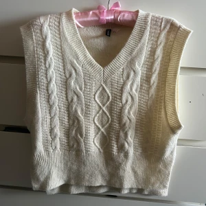 Vit stickad väst från H&M Divided - Säljer en vit stickad väst från H&M Divided i storlek S. Västen har snyggt flätmönster framtill, v-ringad hals och ribbade kanter. Perfekt att styla över en skjorta eller t-shirt för en trendig look. Mjuk och skön kvalitet.