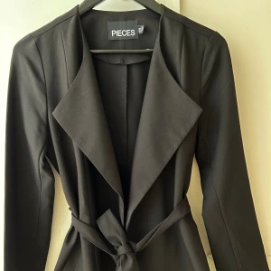 Ny Svart kavajjacka från Pieces - Svart kavajjacka från Pieces med snyggt omlottkrage och bälte i midjan. Jackan har långärmad design och en clean, stilren look som passar till många outfits. Materialet är mjukt och känns skönt mot huden. Perfekt för dig som gillar minimalistisk stil.