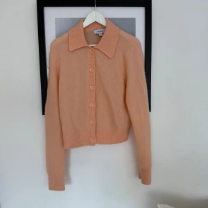 Persikofärgad kofta från & Other Stories - Superfin persikofärgad kofta från & Other Stories med klassisk krage och knappar framtill. Koftan är stickad och har långa ärmar samt ribbade muddar och nederkant. Perfekt för lager-på-lager under kyliga dagar.