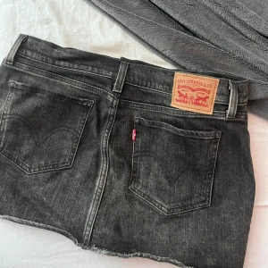 Svart jeanskjol från Levi's, strl 26 - Säljer en svart jeanskjol från Levi's i klassisk femficksmodell. Kjolen har rå kant nertill, bälteshällor och dragkedja med knapp framtill. Perfekt för dig som gillar en avslappnad och cool stil. Storlek 26.