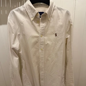 Vit skjorta från Ralph Lauren - Klassisk vit skjorta från Ralph Lauren med broderad logga på bröstet. Skjortan har lång ärm, knappar hela vägen och en snygg krage. Perfekt för dig som gillar stilrena och tidlösa plagg.