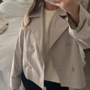 Beige kort trenchcoat med knappar - Snygg kort trenchcoat i beige med dubbelknäppning framtill och stora slag. Jackan har långa ärmar med knappar vid ärmslut och en klassisk, stilren look. Perfekt för dig som gillar minimalistisk stil och vill ha en jacka som funkar till många outfits.
