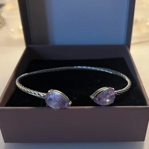 Silverfärgat armband med lila stenar - Snyggt armband i tvinnad silverfärgad metall med två droppformade lila stenar i ändarna. Perfekt accessoar för att ge din outfit lite extra färg och glans.