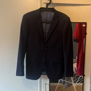 Mörkblå kostym Dressmann slim fit - Snygg mörkblå kostym från Dressmann i slim fit-modell. Kavajen har två knappar, klassiska fickor och slät finish. Byxorna är raka och matchar perfekt till kavajen. Perfekt för dig som vill ha en stilren och modern look. Kostymen i bra skick, kavaj använd ett 10-tal gånger och därav ett superfint skick. Byxorna har använts separat mer och har lite tecken på användning. Främst sömmen på ett av benen som lossnat lite men inget som skadar användningen. Nypris mellan 2000-2500:- mitt pris: 699