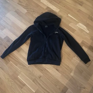 Svart Peak Performance hoodie - Svart hoodie från Peak Performance med dragkedja och broderad logga på bröstet. Tröjan har huva, långa ärmar och är gjord i mjuk bomullsmix. Perfekt för chill dagar eller när du vill ha en enkel och stilren look. Storlek 170 / XS-S