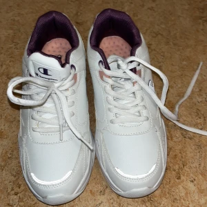 Vita sneakers från Champion - Snygga vita sneakers från Champion med lila innerfoder och rosa prickig innersula. Skorna har klassisk snörning och är tillverkade i syntetmaterial med detaljer i textil. Perfekta för dig som gillar sportig och fräsch stil.