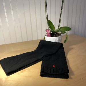 Svart stickad halsduk Polo Ralph Lauren - Svart halsduk från Polo Ralph Lauren med klassisk röd broderad logga. Stickad i mjukt material med ribbade kanter för extra stilpoäng. Perfekt längd för att vira flera varv och ger en clean look till din outfit.