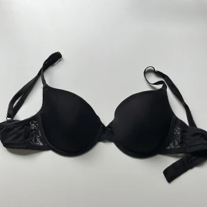 Svart spetsdetaljerad bh från Twilfit 70D - Snygg svart bh från Twilfit med vadderade kupor och fina spetsdetaljer på sidorna. Axelbanden är justerbara och modellen ger ett stilrent och klassiskt intryck. Perfekt för dig som gillar en enkel men ändå elegant look.